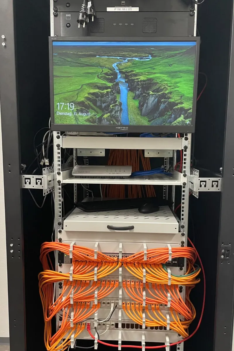 Offener Serverschrank mit Monitor, Router und sauber strukturierter orangefarbener Verkabelung.